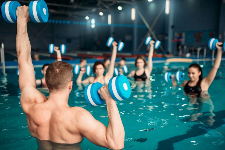 En grupp personer deltar i en gruppträning i en simbassäng inomhus ledda av en instruktör. I sina händer håller de en form av hantel som de lyfter upp i luften.