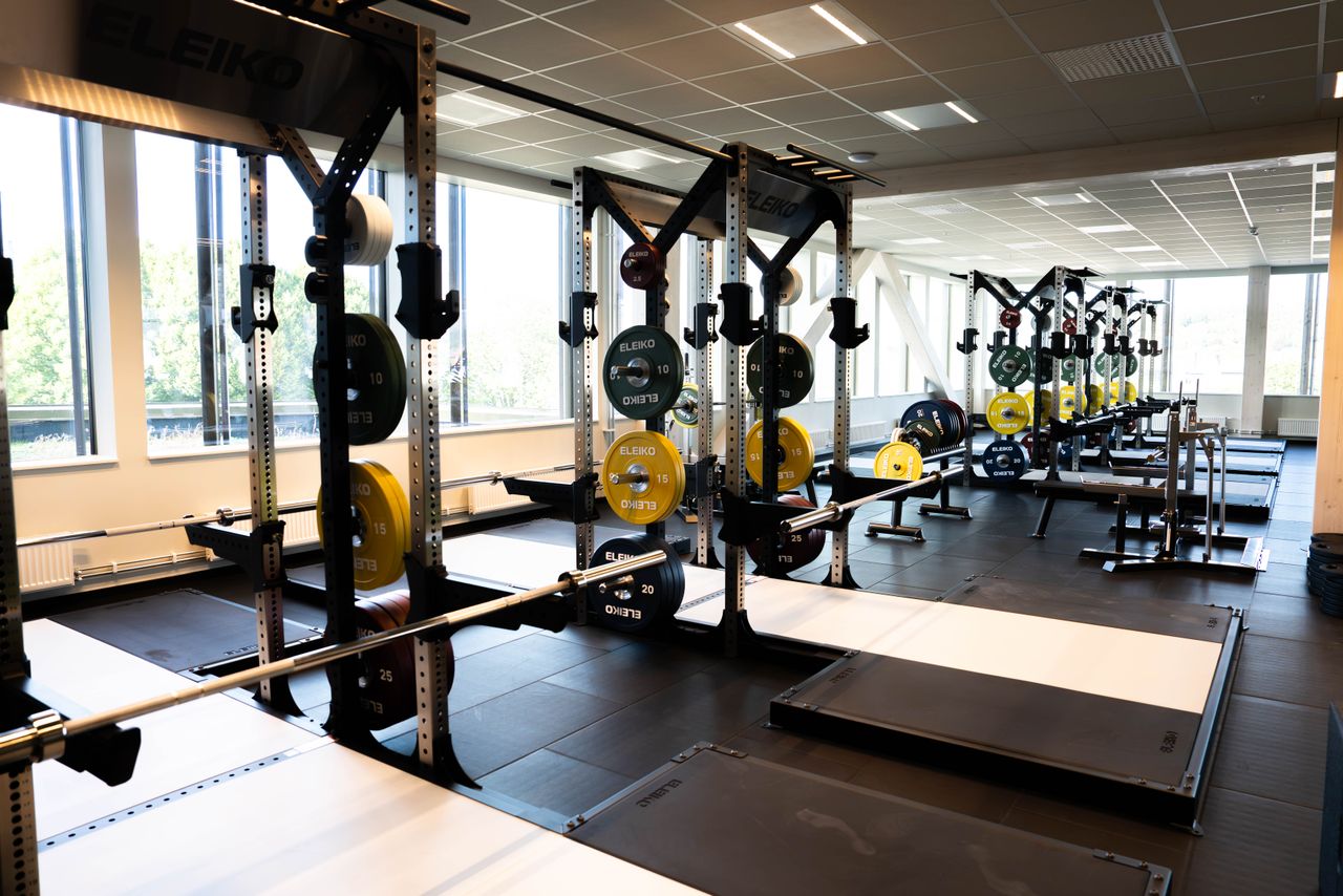 Ett ljust gym med flera knäböjställningar, skivstänger och viktskivor som är prydligt uppställda. Stora fönster släpper in naturligt ljus, och golvet har lyftplattformar. Gymmet ser rent och tomt ut.