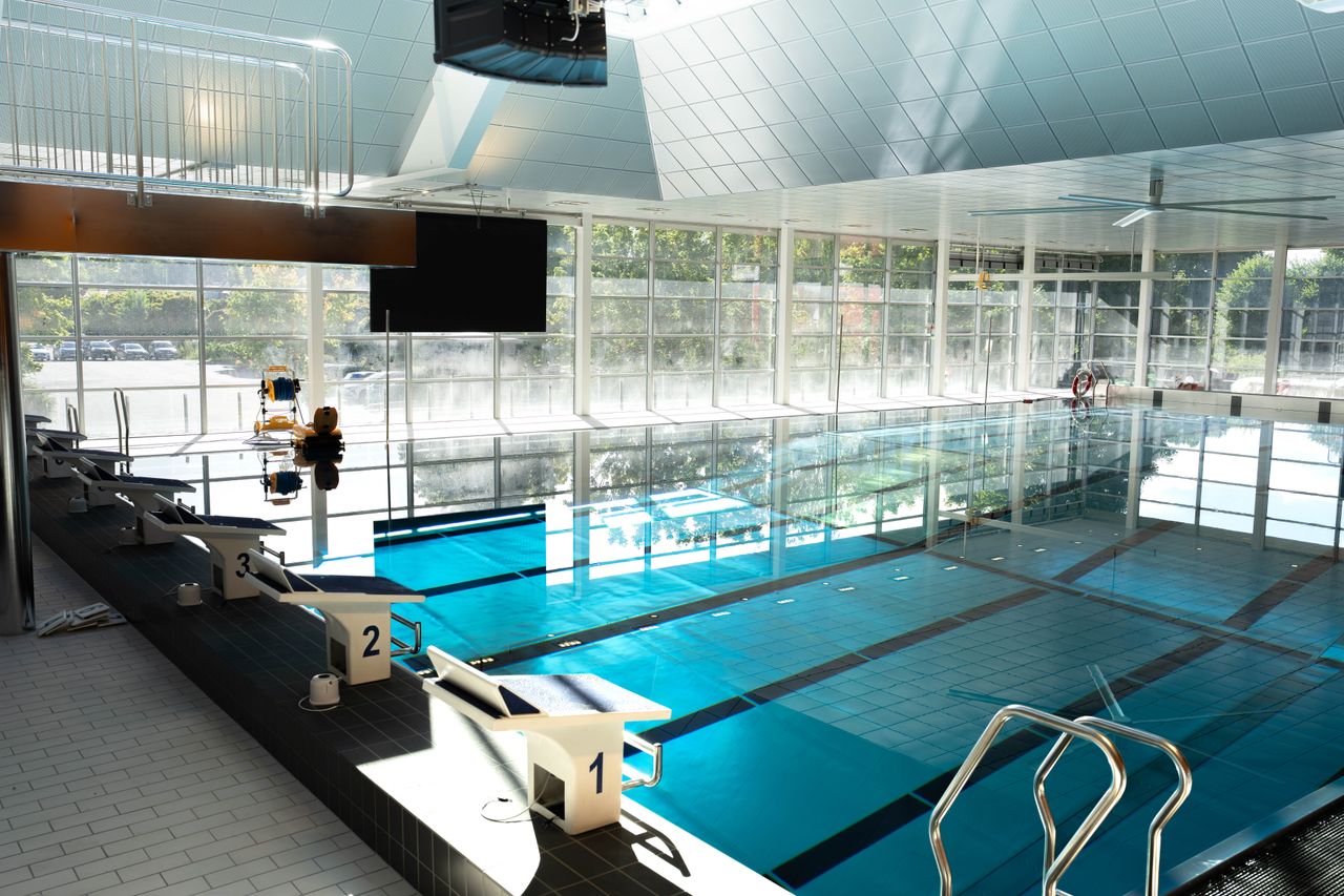 En inomhuspool med fem startblock, klart vatten, stora fönster som släpper in solljus och en badvakt som sitter vid poolkanten.