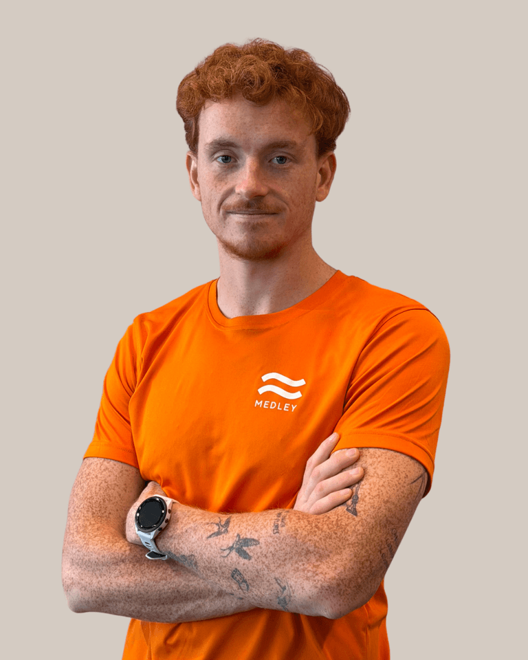 En person med rött hår och fräknar, klädd i en orange Medley-t-shirt och med en sportklocka, som står med armarna i kors.