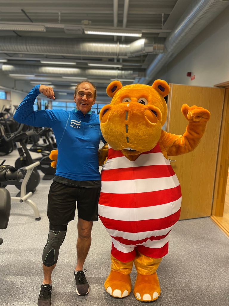 En leende man med en benprotes böjer armen medan han står bredvid en person i en orange flodhästdräkt med röda och vita ränder inne i ett gym.