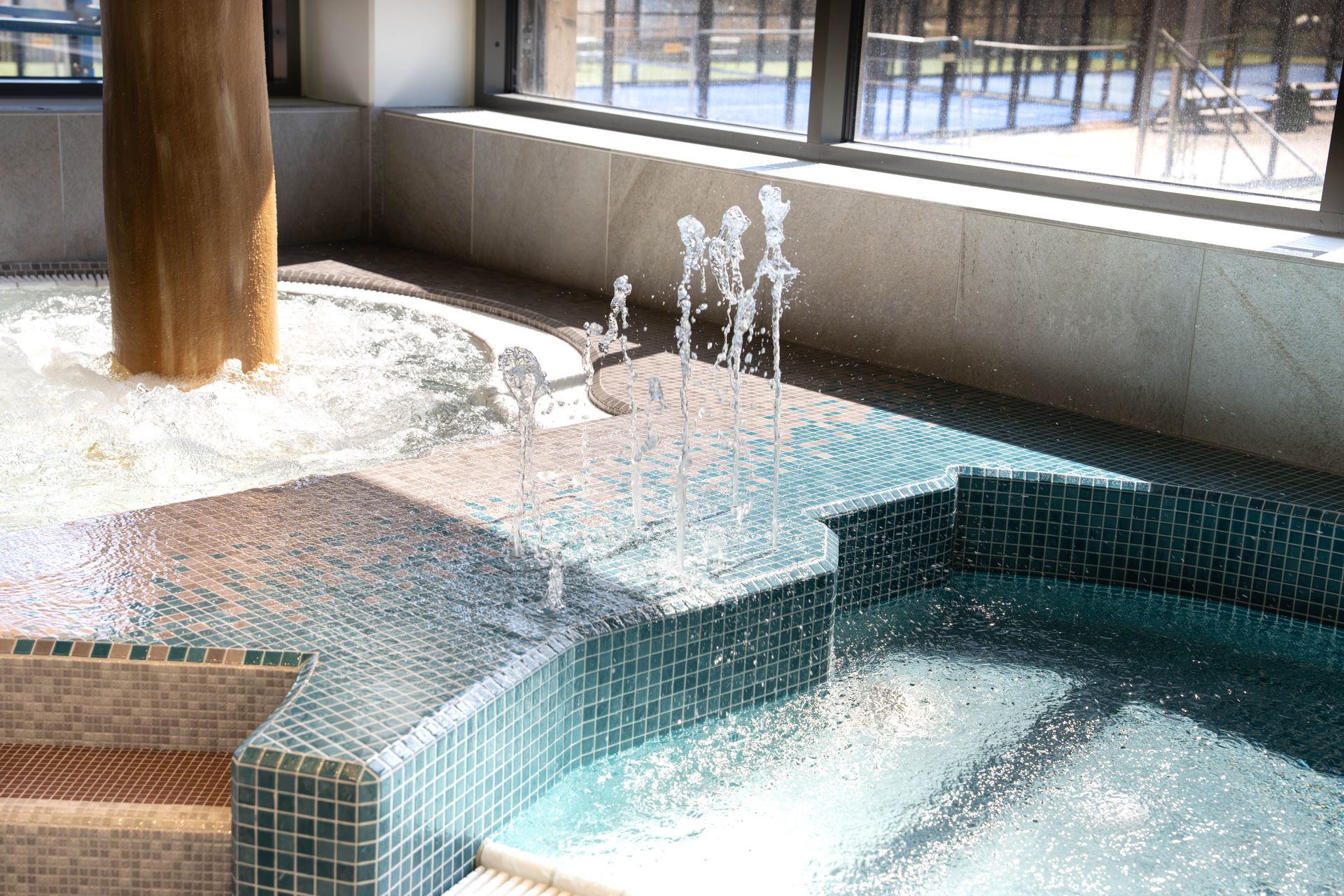 Inomhuspoolområde med turkosa kakelplattor, en bubblande jacuzzi, flera små vattenfontäner och solljus som strömmar in genom stora fönster och speglas i vattnet. Utanför fönstren syns tennisbanor.