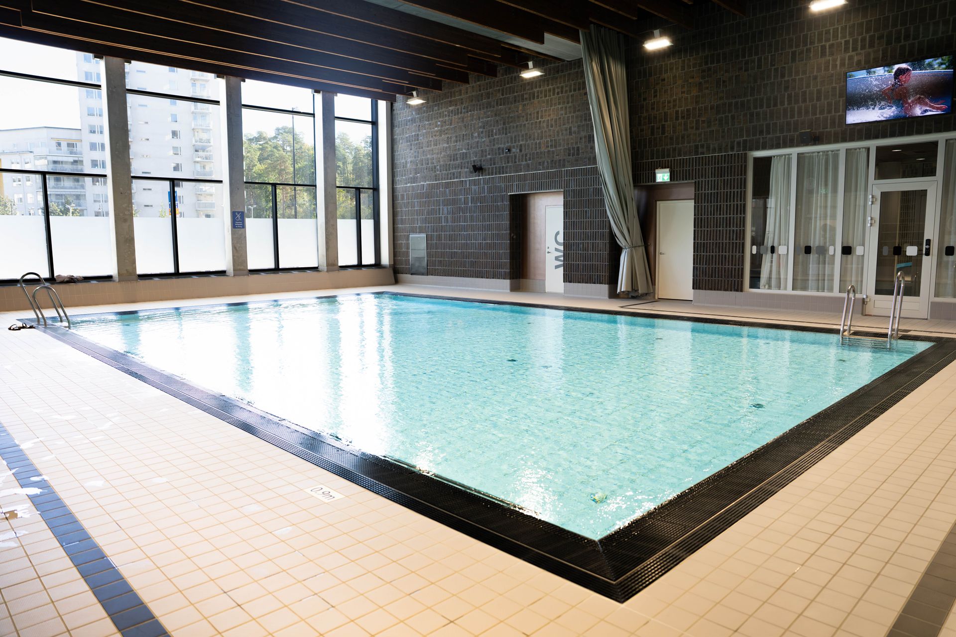 En inomhuspool med klart vatten, omgiven av kaklat golv, stora fönster och grå tegelväggar. Det finns stegar i två av poolens hörn och en fördragen gardin på ena sidan.