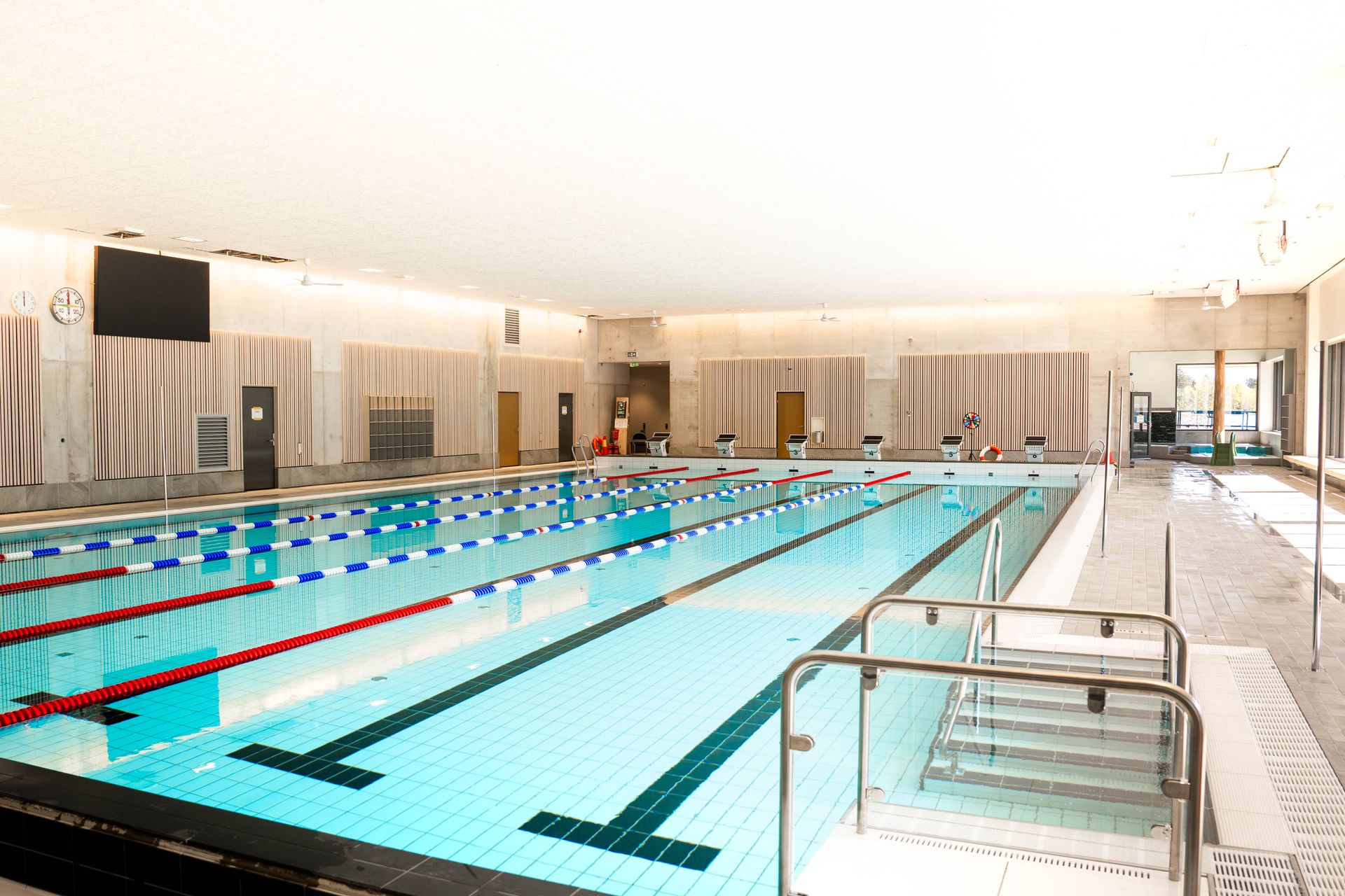Inomhuspool med klart blått vatten, banavgränsningar, startblock och en livräddningsstol. Poolområdet är väl upplyst av naturligt ljus som strömmar in genom stora fönster till höger. I förgrunden syns en trappa som leder ner i poolen.