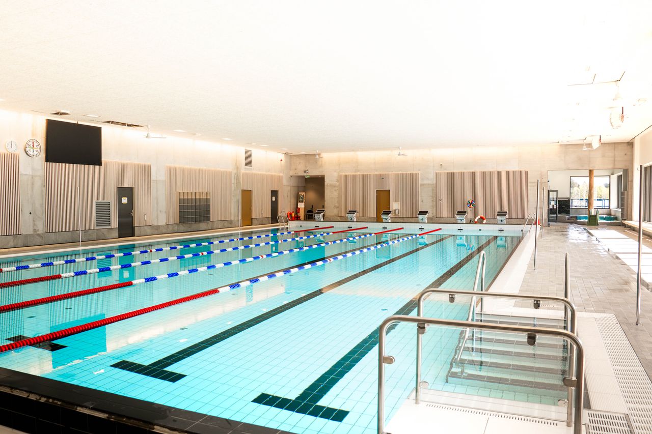 Inomhuspool med klart blått vatten, banavgränsningar, startblock och en livräddningsstol. Poolområdet är väl upplyst av naturligt ljus som strömmar in genom stora fönster till höger. I förgrunden syns en trappa som leder ner i poolen.
