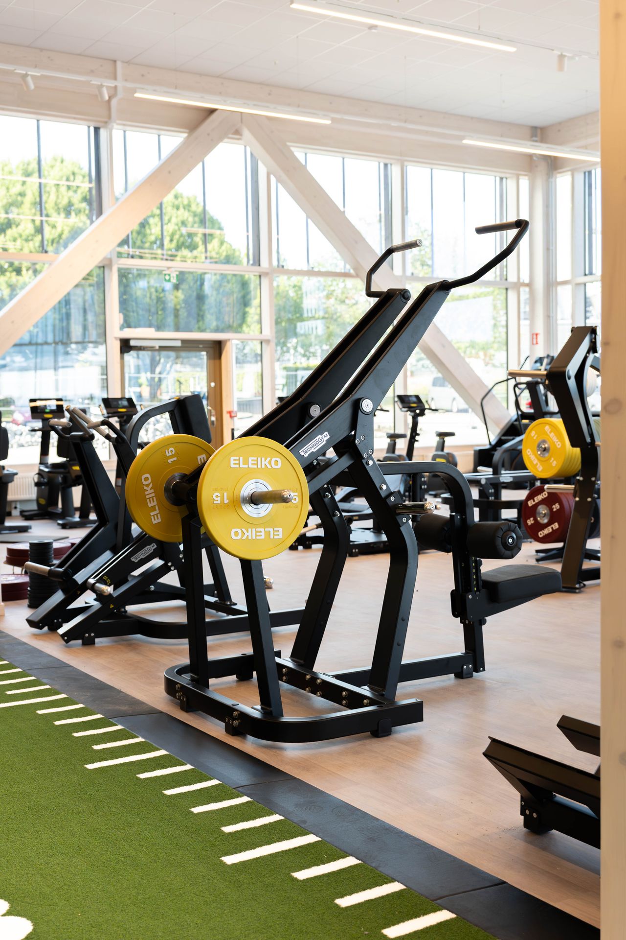Ett gym med styrketräningsmaskiner, skivstänger med gula ELEIKO-vikter på 15 kg och stora fönster som släpper in naturligt ljus. I förgrunden syns grön konstgräsmatta.