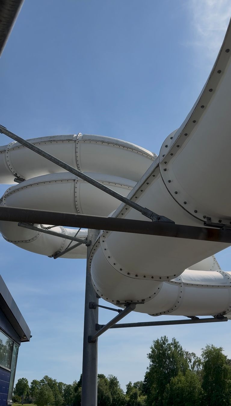 Stora vita, slutna vattenrutschbanor slingrar sig ovanför marken på metallstöd mot en blå himmel, med gröna träd synliga i bakgrunden.