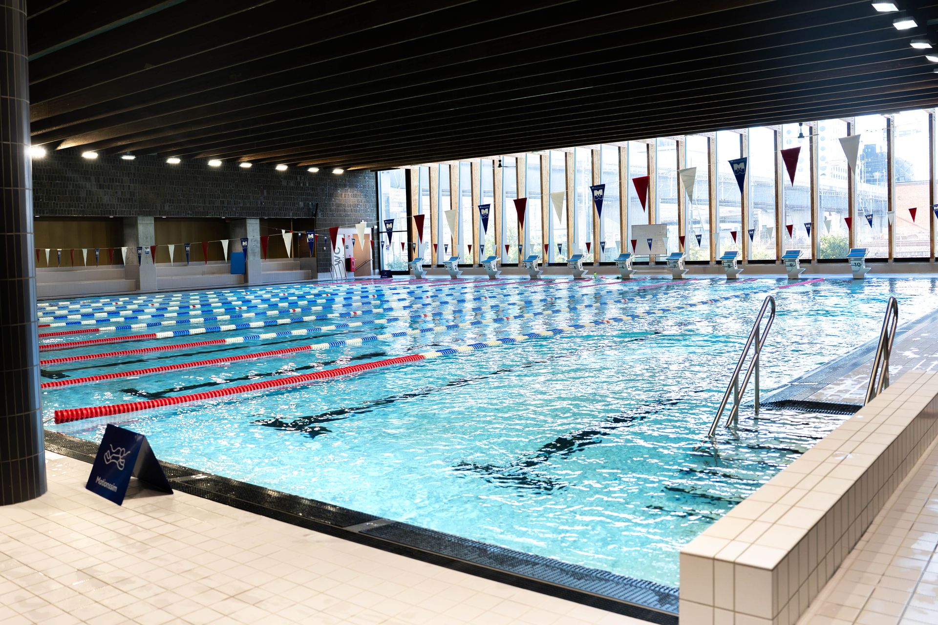 Inomhuspool med flera banor, banavdelare i rött, blått och vitt, en hopptorn, startblock och stora fönster som släpper in naturligt ljus. En stege vid poolen och en skylt med texten ”Hoppning förbjuden” syns tydligt.