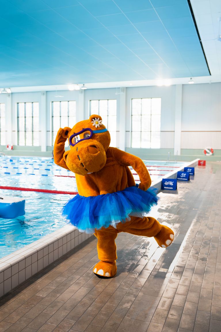 En maskot i en brun flodhästdräkt, med blå simglasögon och en blå tutu, poserar vid en inomhuspool.