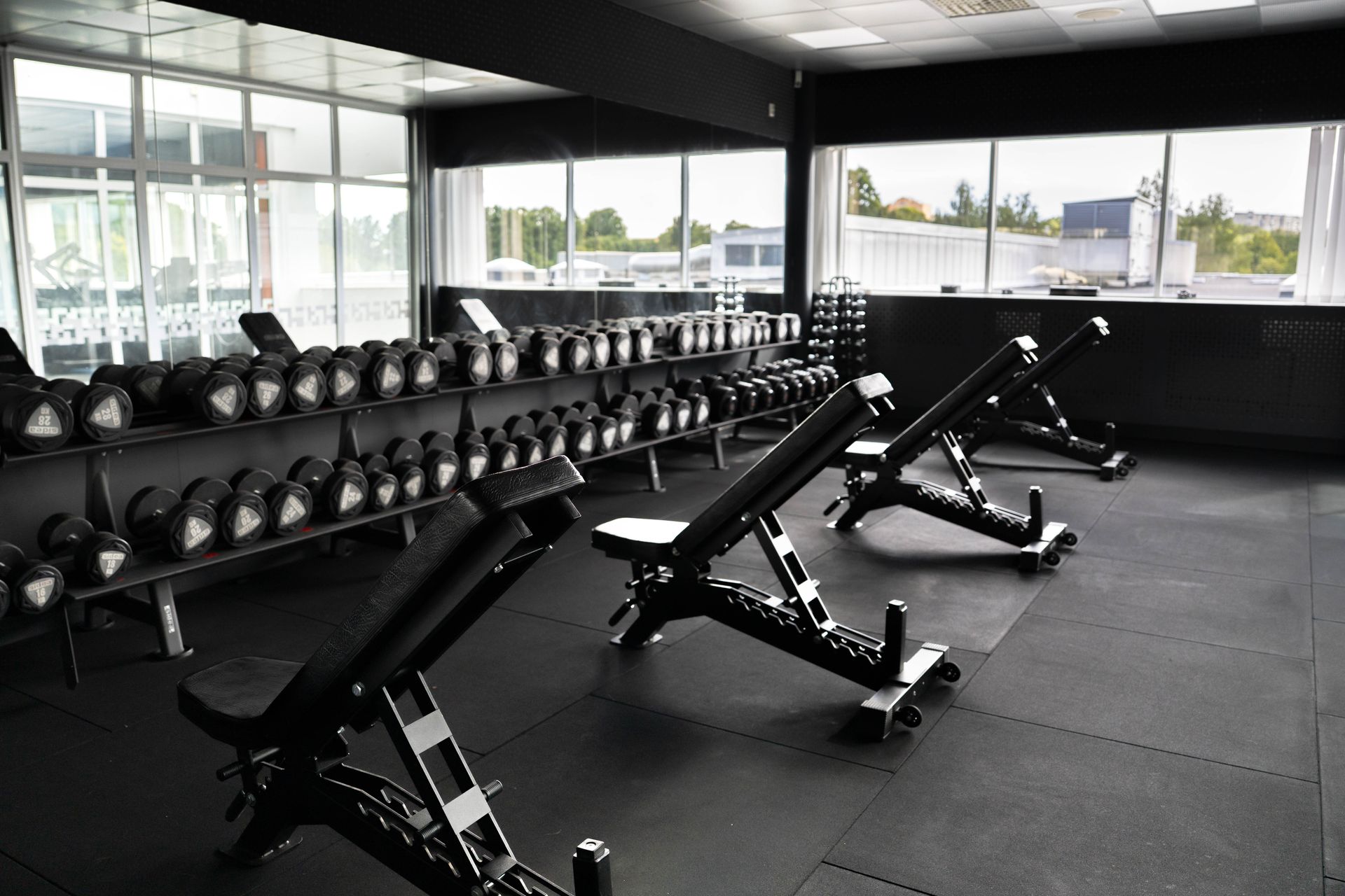 Ett modernt gym med justerbara bänkar och flera ställningar med hantlar, beläget vid stora fönster som släpper in naturligt ljus. Lokalen är ren, öppen och inredd för styrketräning.