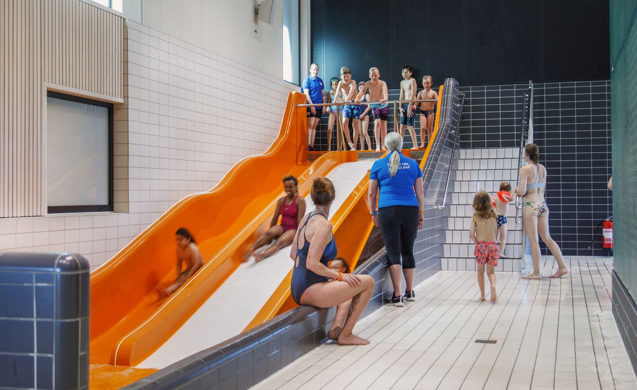 Barn som leker i en stor orange vattenrutschbana inomhus, medan andra väntar på trappan och vid poolkanten.