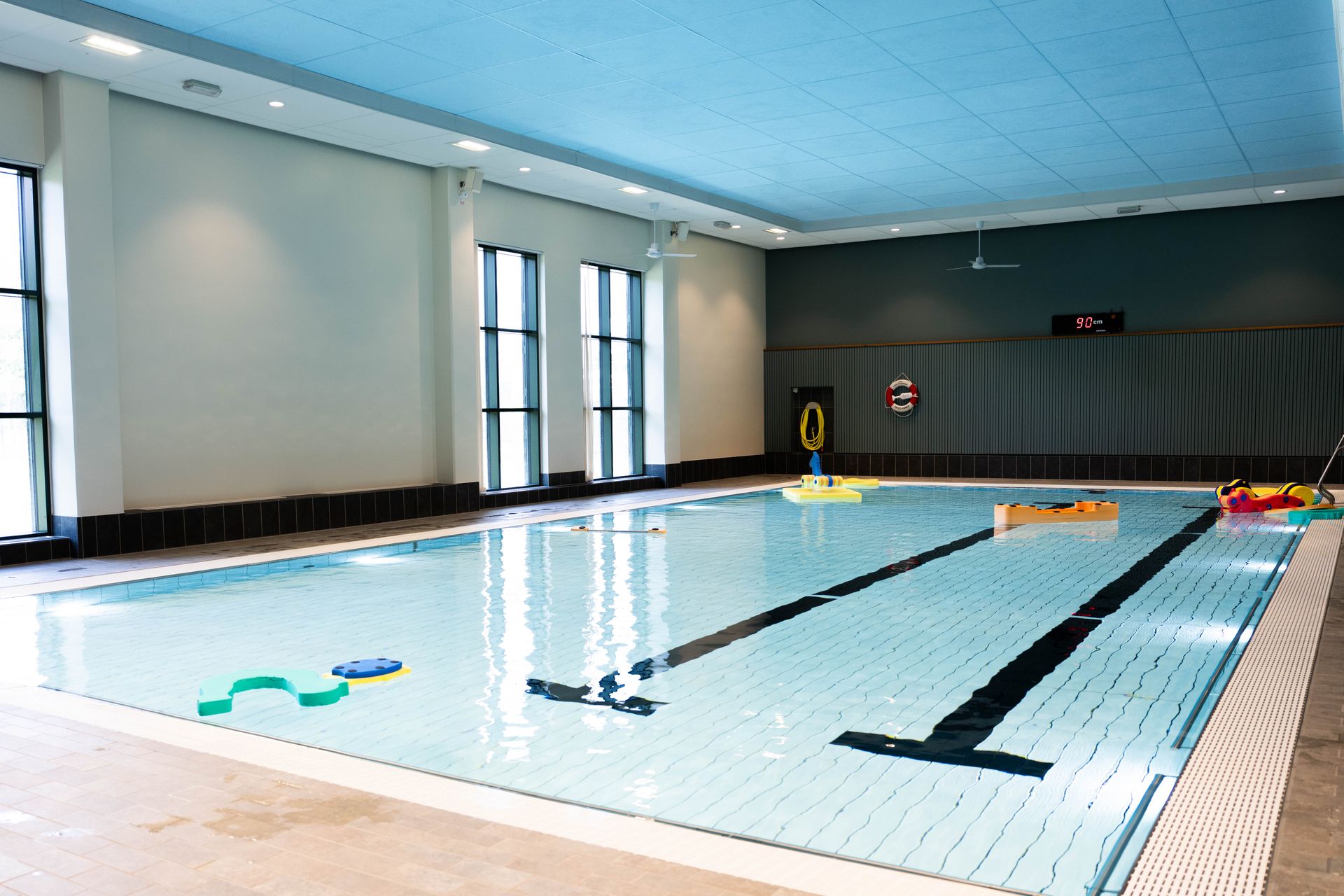 Inomhuspool med stora fönster, badleksaker som flyter i vattnet och en digital klocka på bakväggen. Poolområdet är väl upplyst och tomt på människor.