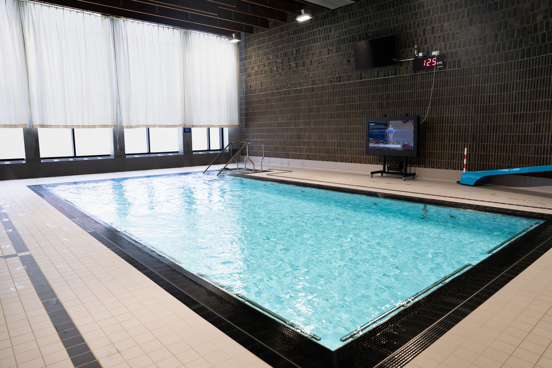 Inomhuspool med klart vatten, kaklat område runt poolen, en blå rutschkana, ledstänger, stora fönster med tunna gardiner samt en väggmonterad TV och en digital klocka som visar 1:25. Det finns inga människor i bild.