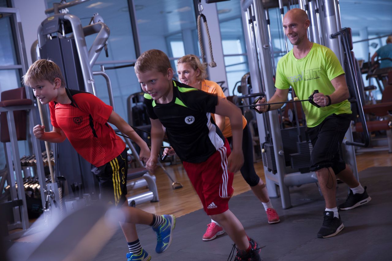 Bild på en mamma och pappa som tränar med två söner inne på gymmet, föräldrarna agerar motstånd med gummiband medans barnen springer framåt