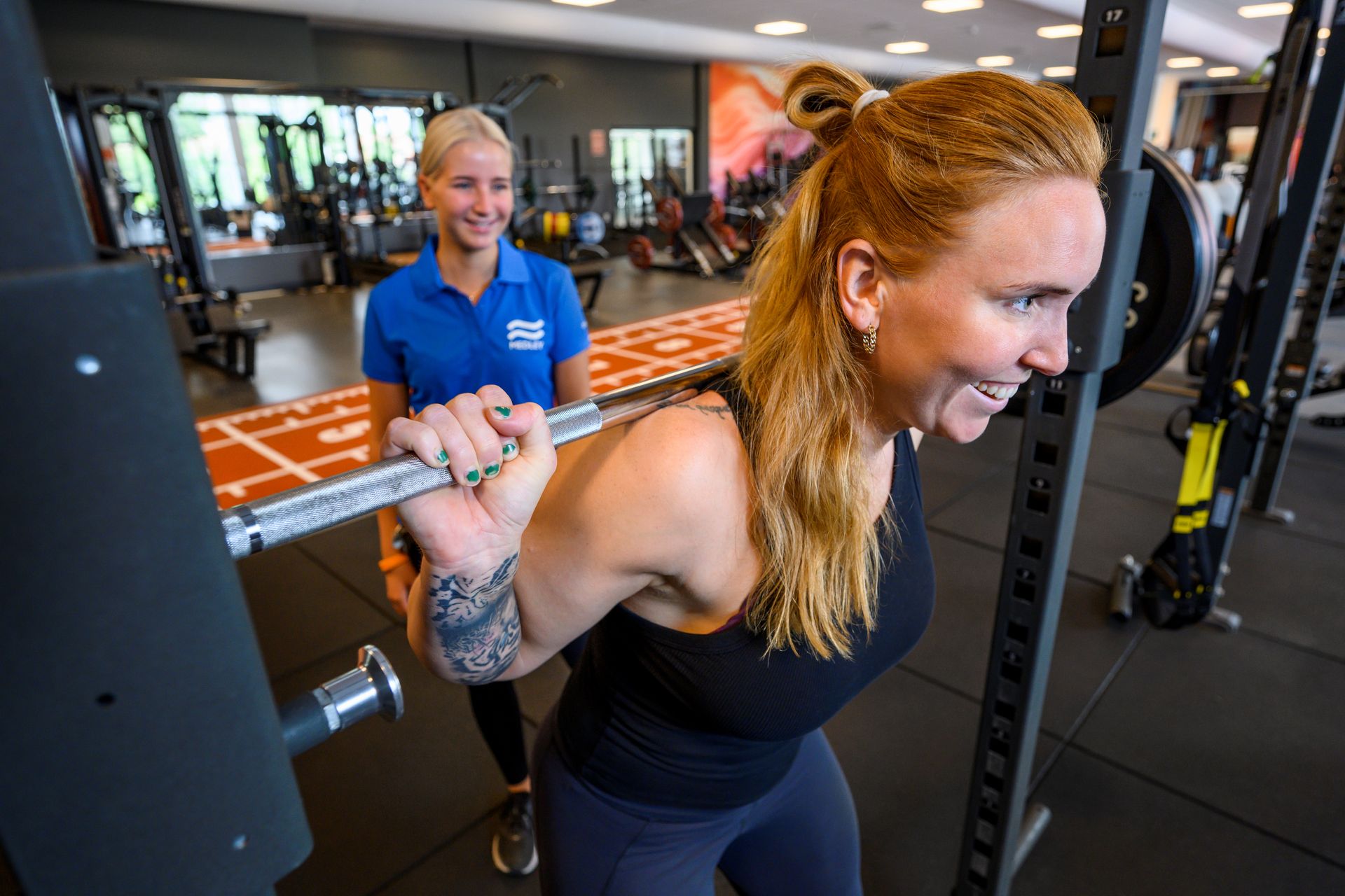 En person som ler och är klädd i ett svart linne lyfter en skidstång på ett gym. En tränare klädd i blått står bakom och ser till att lyftet sker korrekt.