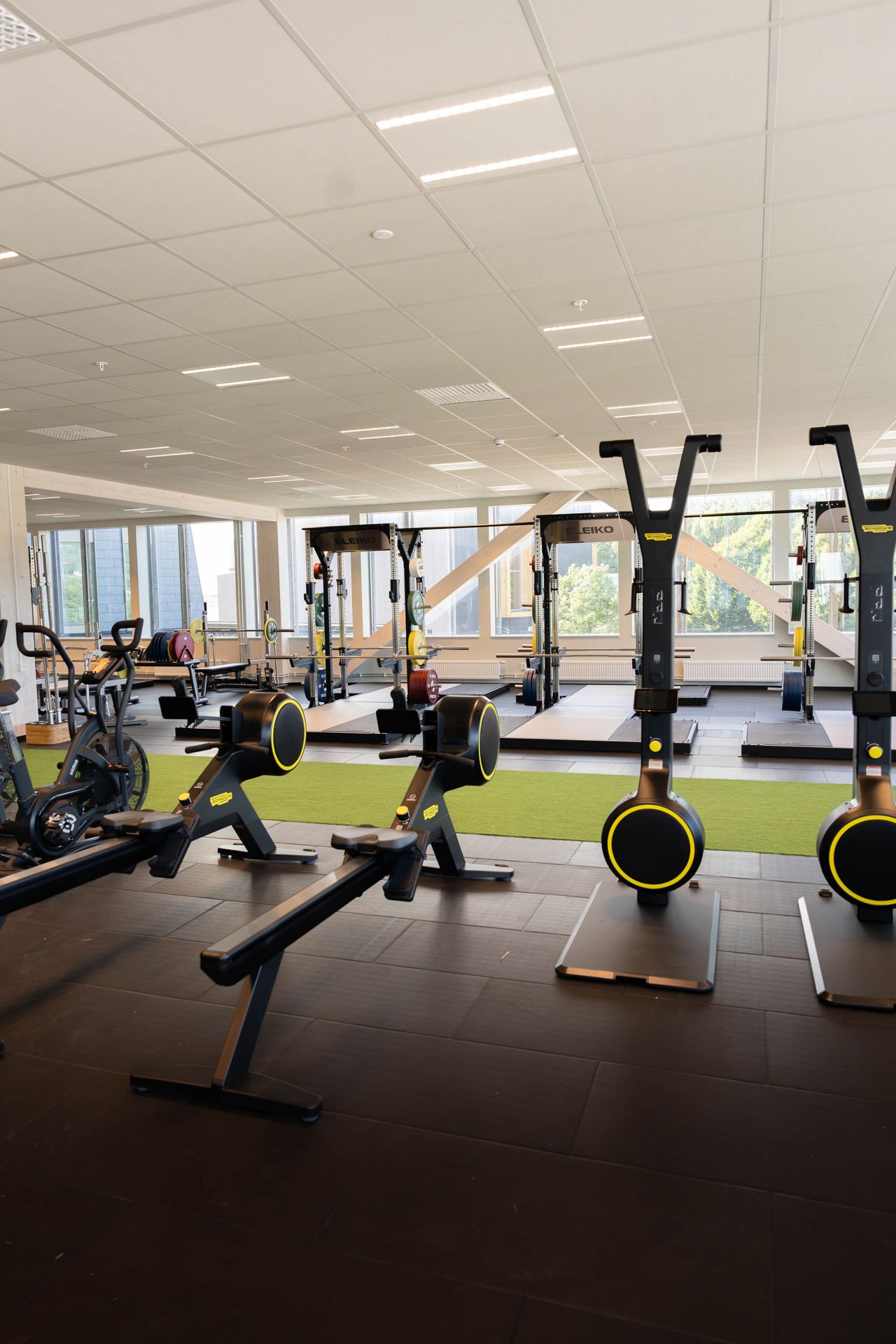Ett modernt gym med roddmaskiner, skidergometer och styrketräningsplattformar. Ett ljust utrymme med stora fönster, grönt konstgräs och olika träningsredskap. I bakgrunden syns några personer som tränar.