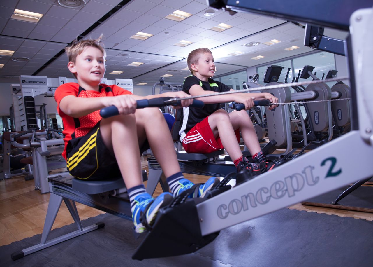 Två unga killar sitter bredvid varandra i roddmaskiner och drar för glatta livet