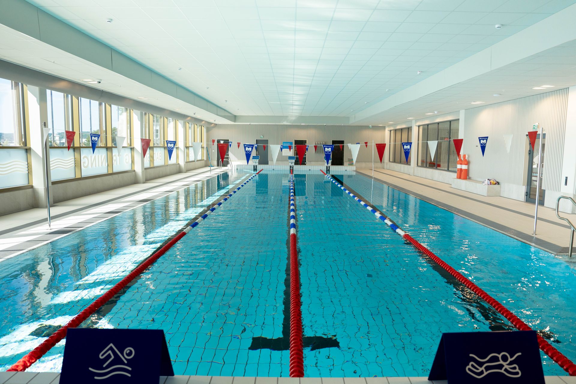 Inomhuspool med klart blått vatten, banavgränsningar och solljus som strömmar in genom stora fönster. Skyltar och flaggor som markerar säkerhetsreglerna syns längs sidorna, vilket skapar en ren och välordnad atmosfär.
