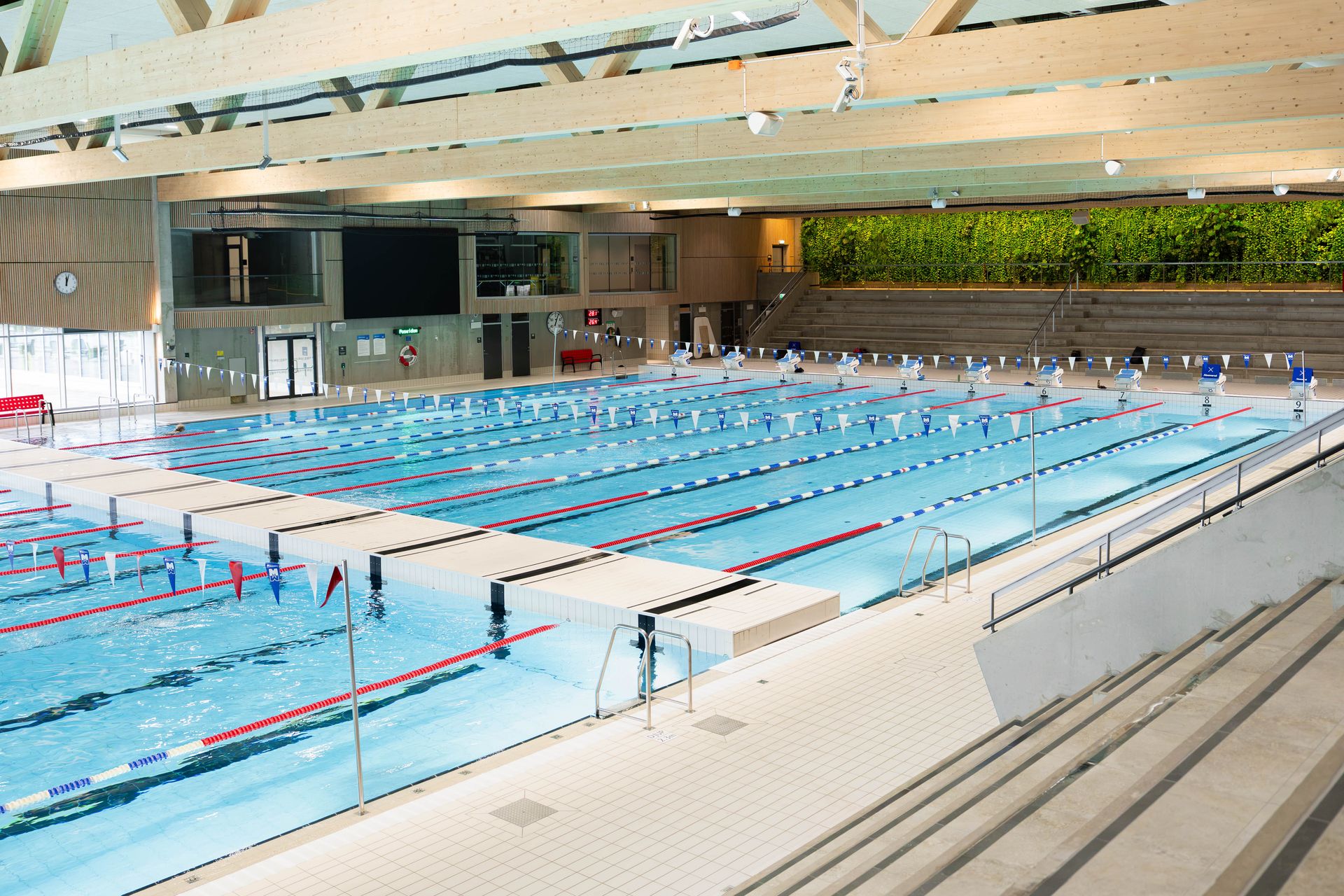 En inomhuspool med flera banor, hopptorn, banmarkeringar och åskådarplatser. Lokalen är väl upplyst av naturligt ljus och har en modern design med träbjälkar.