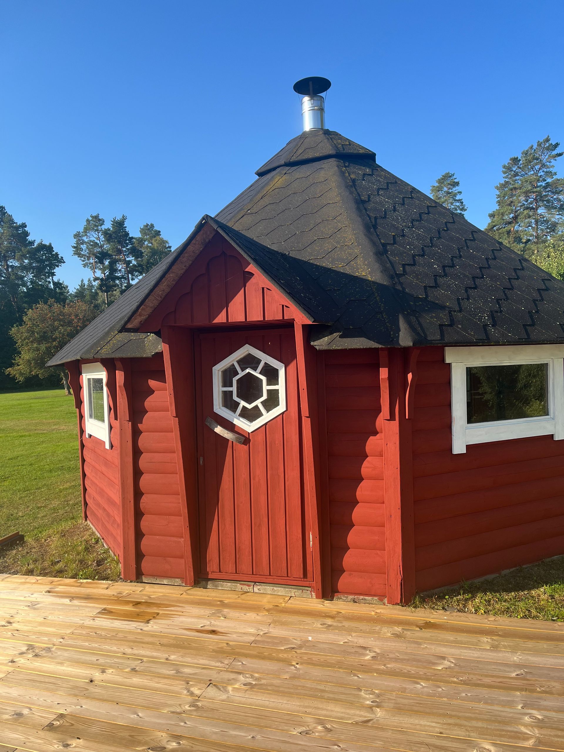 En liten röd trästuga med sexkantig form, fönster med vita ramar och en skorsten på det mörka taket står på en träterrass med gräs och träd i bakgrunden under en klarblå himmel.