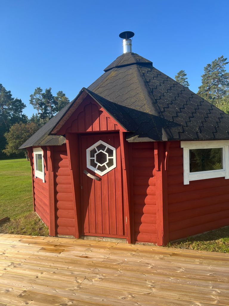 En liten röd trästuga med sexkantig form, fönster med vita ramar och en skorsten på det mörka taket står på en träterrass med gräs och träd i bakgrunden under en klarblå himmel.
