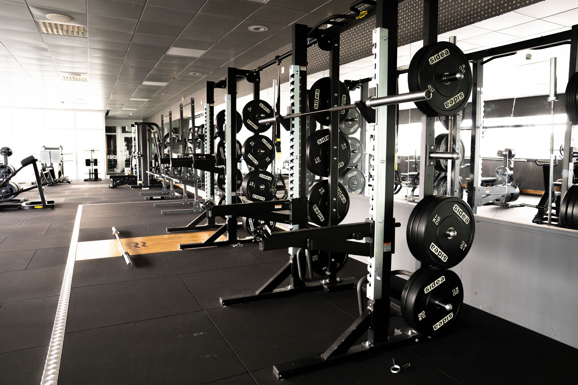 Ett tomt gym med styrketräningsställ, skivstänger och viktskivor uppradade längs en spegelvägg; träningsmaskiner och bänkar syns i bakgrunden.