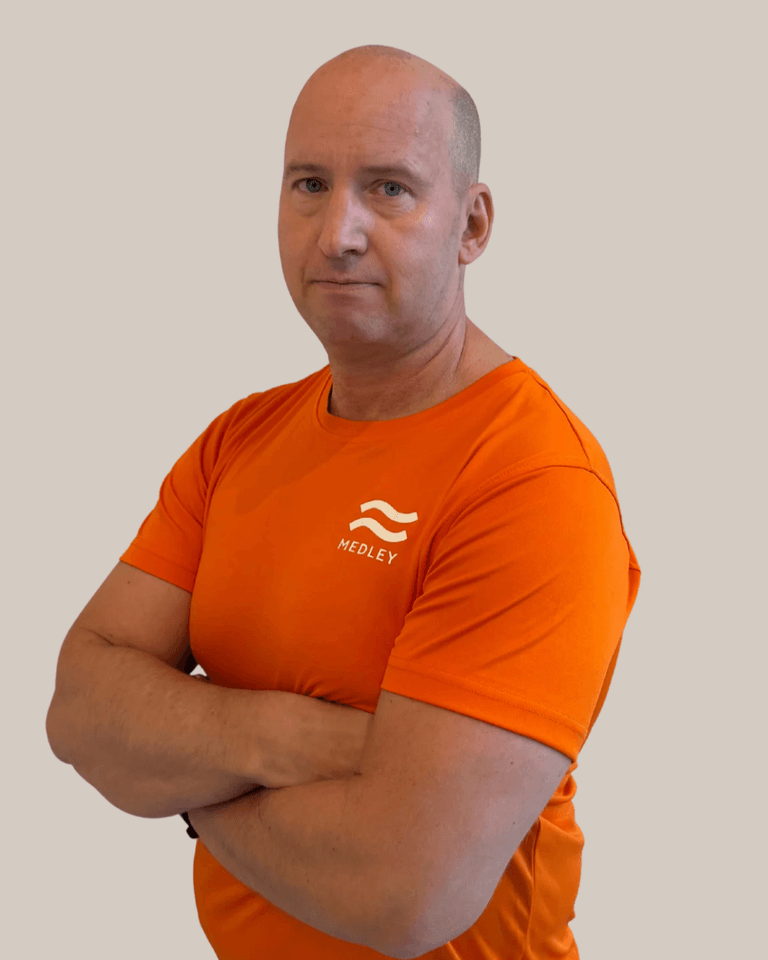 En skallig man med korslagda armar, klädd i en orange T-shirt med Medley-logotypen.