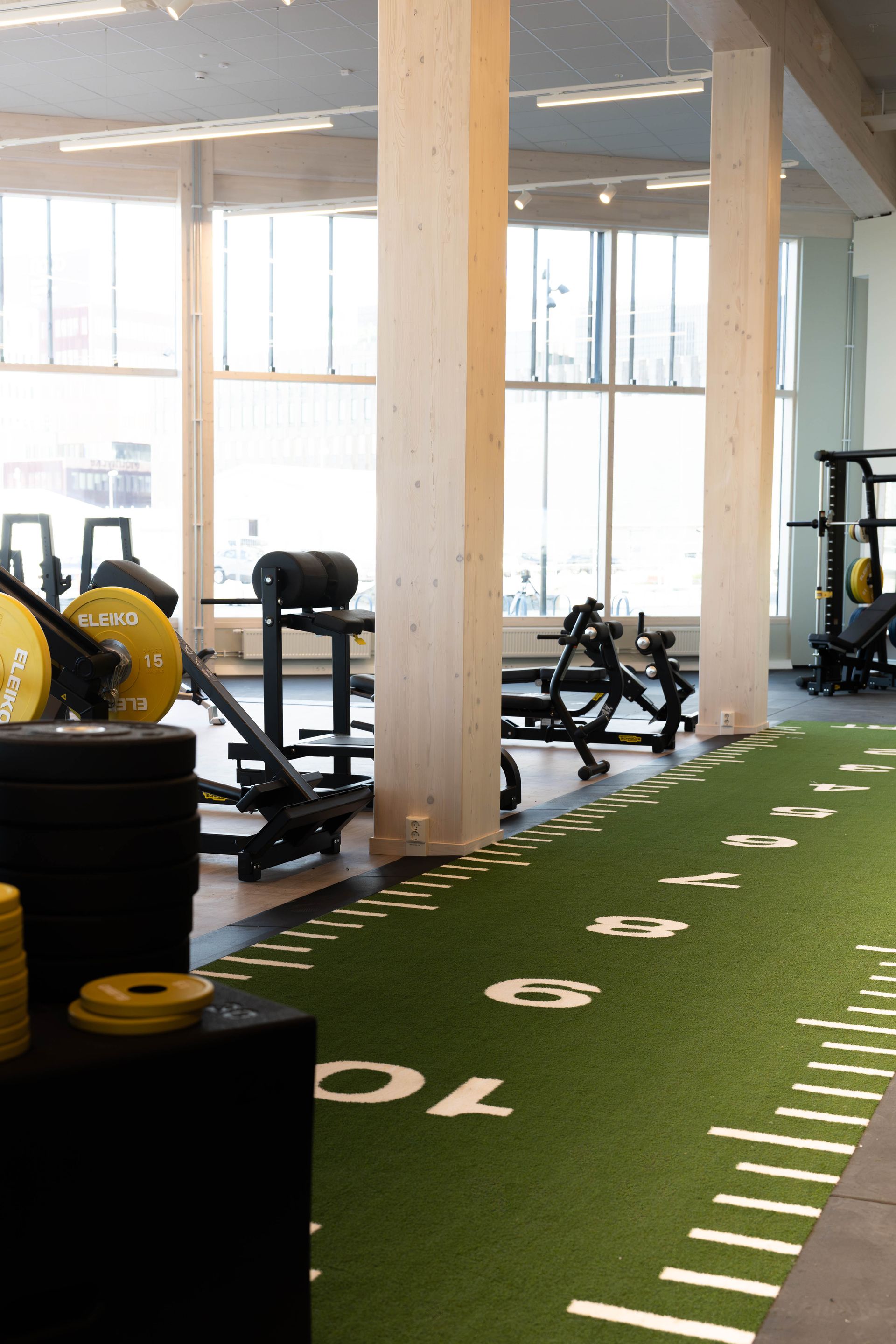 Ett gym med styrketräningsmaskiner, gula viktskivor och en grön konstgräsyta markerad med vita siffror, ungefär som en fotbollsplan. Stora fönster släpper in naturligt ljus.