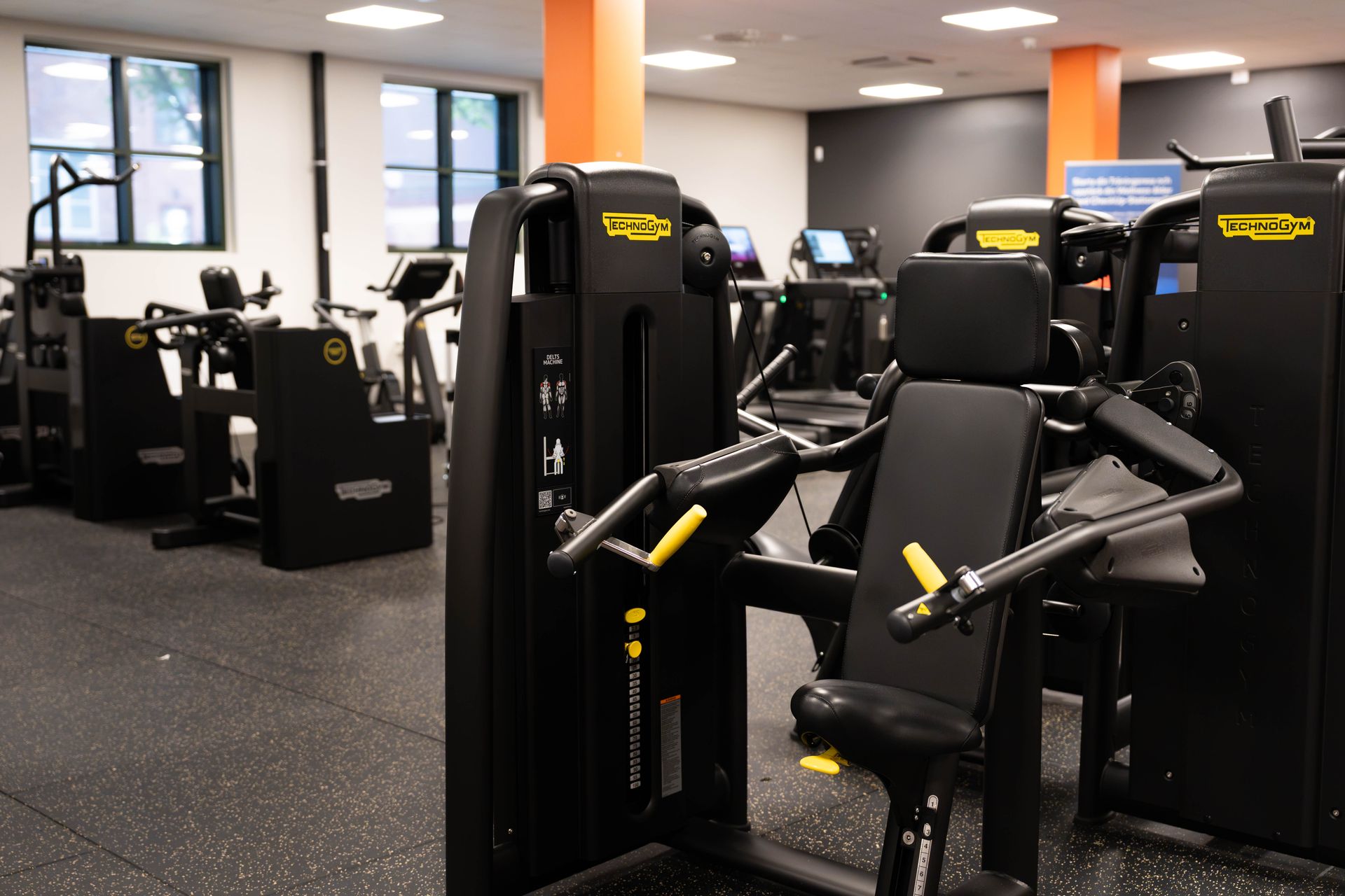 Ett rent och modernt gym med olika träningsmaskiner från Technogym, svart golv, klarorange pelare och stora fönster som släpper in naturligt ljus.