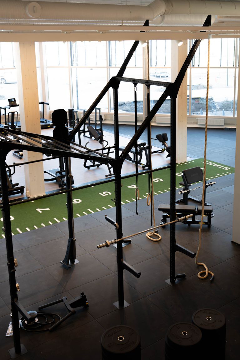 Ett modernt gym med svarta träningsmaskiner, en grön konstgräsbana med nummermarkeringar, skivstänger och ett klätterrep – allt i ett ljust rum med stora fönster som släpper in naturligt ljus.