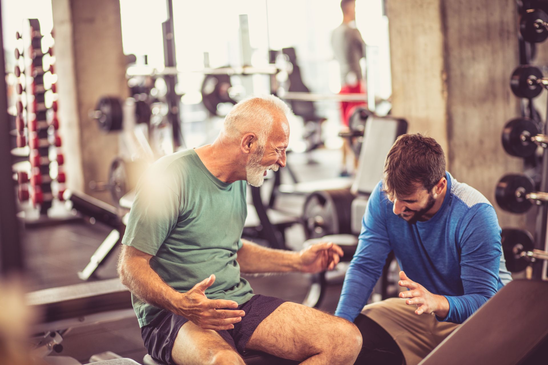 En man i träningskläder sitter på en bänk och får tips och instruktioner av en tränare på ett gym.