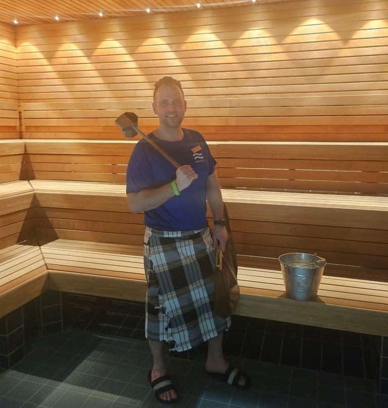 En man i en bastu, klädd i en blå t-shirt och en rutig kilt, som håller en träslev bredvid en vattenhink av metall.