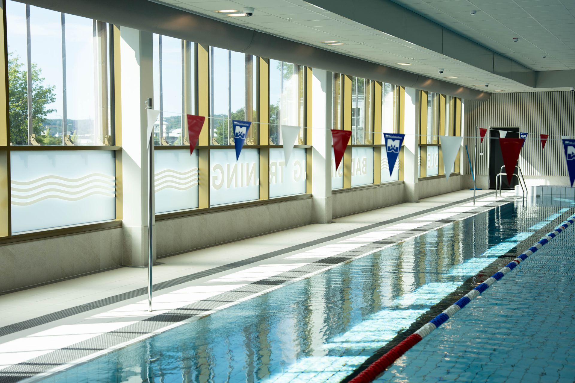En inomhuspool med klart vatten, röda och blå banavgränsare, stora fönster som släpper in solljus och röda triangulära flaggor som hänger ovanför poolen.
