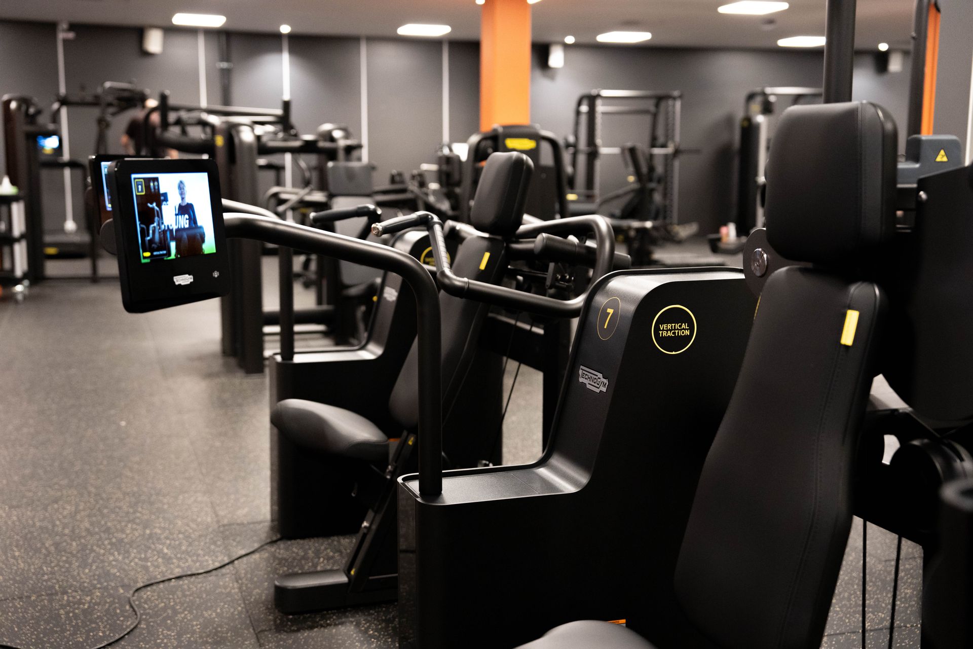 En rad svarta vertikala dragträningsmaskiner av märket Technogym i ett ljust, modernt gym med mörkgrå väggar och orange pelare. En maskin i förgrunden har en skärm som visar en träningsguide.