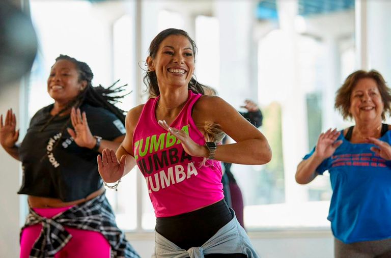 Tre personer deltar i en gruppträning där man dansar. Deltagarna ser glada ut och har färgglada träningskläder på sig. På en av tröjorna står det Zumba i kulörta färger.