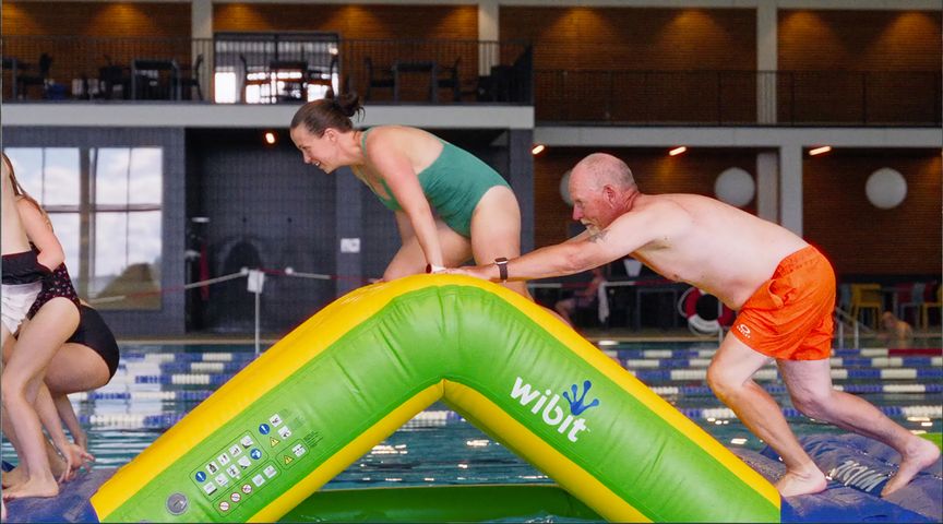 Två vuxna klättrar på ett gult och grönt uppblåsbart hinder i en inomhuspool, medan de ler och har roligt. I bakgrunden syns simmare och sittplatser.