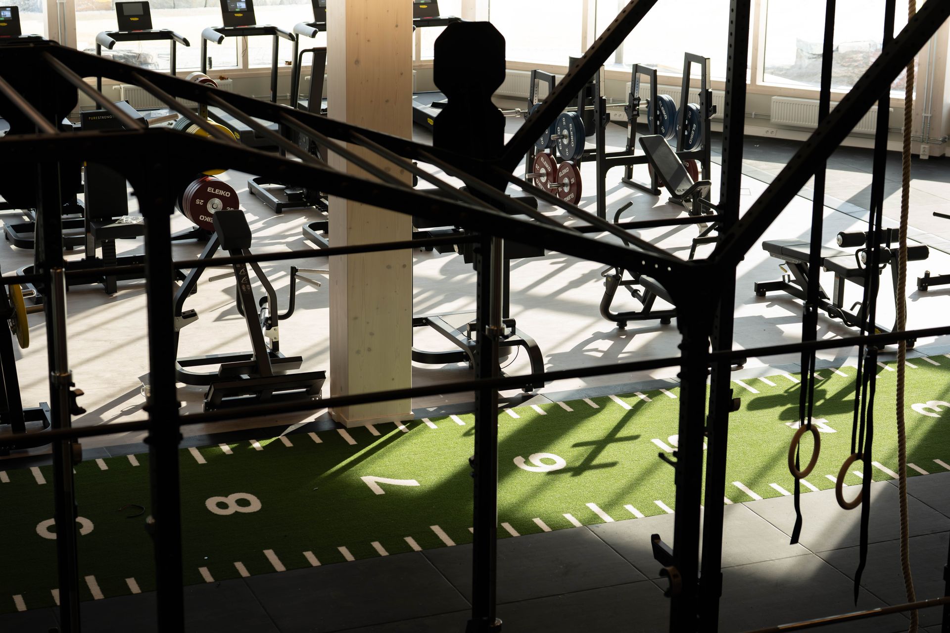 Ett gym med styrketräningsmaskiner, bänkar och löpband. I förgrunden syns en grön gräsmatta med vita siffror samt diverse träningsredskap, bland annat gymnastikringar och stänger. Solljuset strömmar in genom stora fönster.