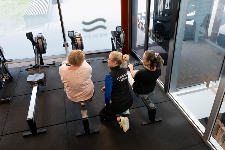 Tre kvinnor befinner sig på ett gym; två sitter vid roddmaskinerna, medan den tredje, klädd i en personlig tränarväst, knäböjer mellan dem och ger dem råd. Stora fönster släpper in naturligt ljus.