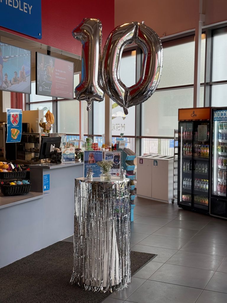Ett bord med silverfärgad frans, dekorerat med tio stora silverfärgade ballonger, står uppställt i en snackbar med drycker, snacks och ett kylskåp i bakgrunden, för att fira ett tioårsjubileum eller ett annat evenemang.