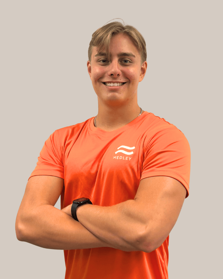 En leende ung man med korsade armar, klädd i en orange sport-T-shirt med ordet Medley tryckt på bröstet.