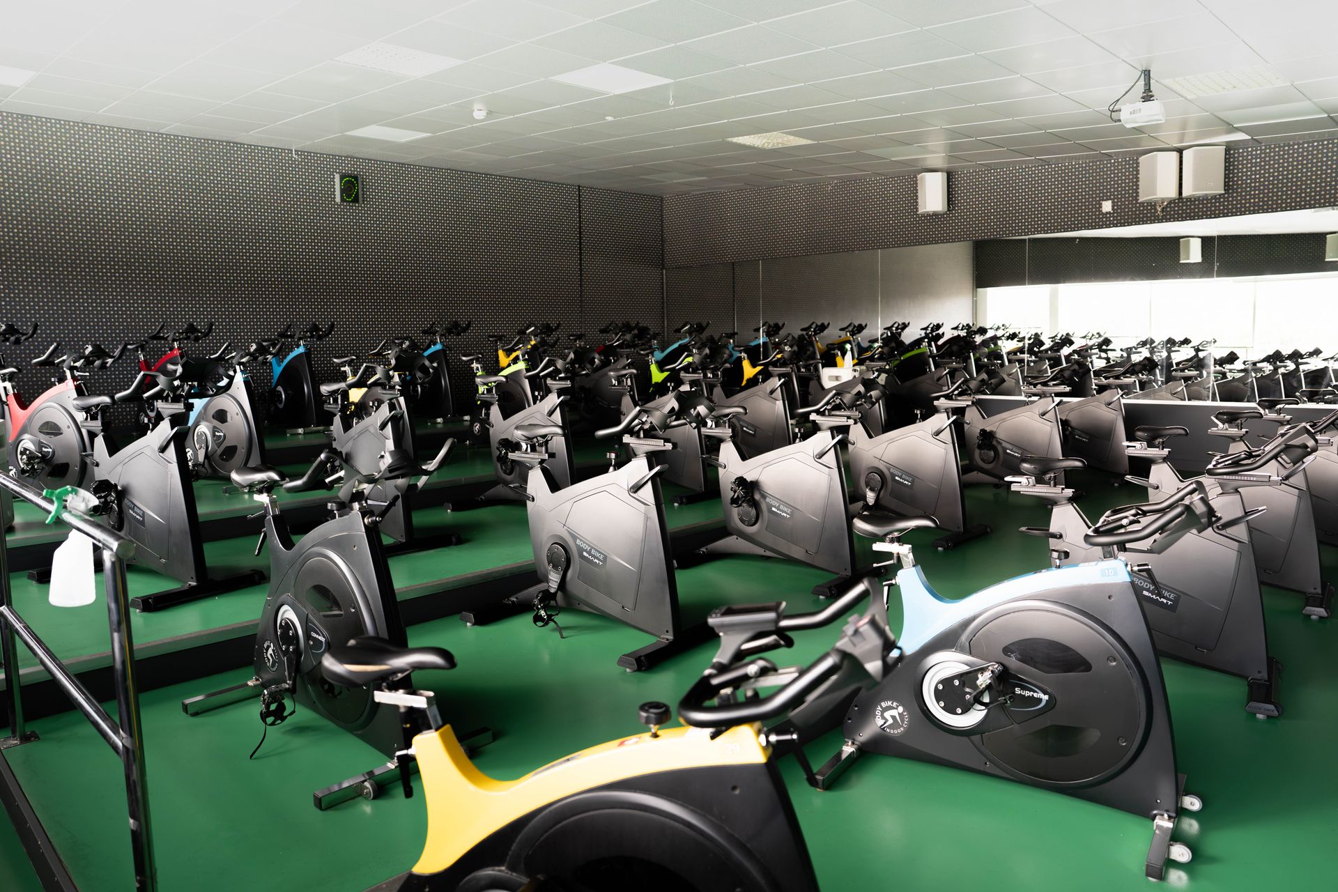 Ett gym med rader av motionscyklar på ett grönt golv, vända mot en spegelvägg. Motionscyklarna har olika färger, och rummet är väl upplyst av taklampor.