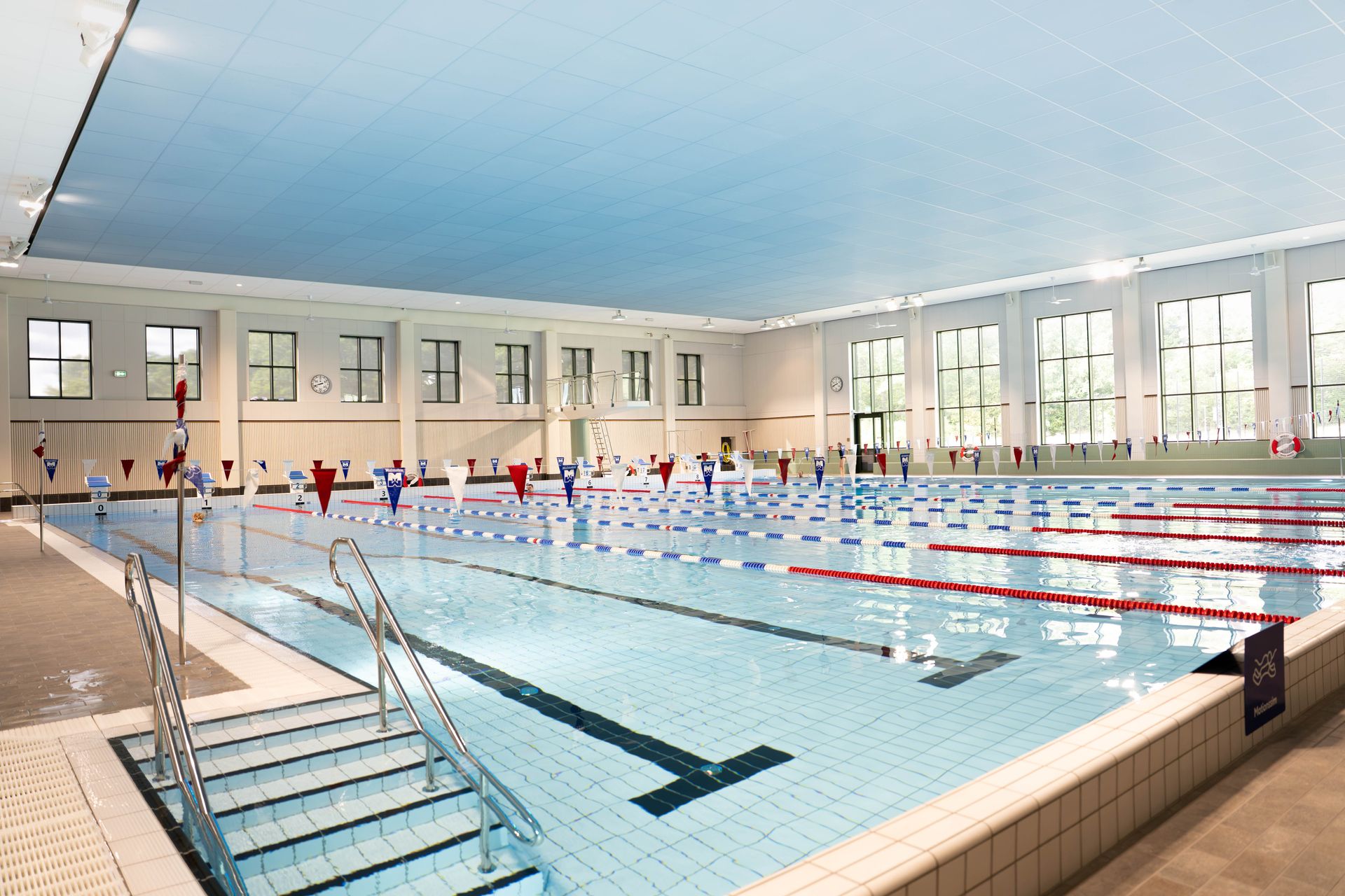 Inomhuspool med flera banor, blått vatten och banavgränsare. Stora fönster sträcker sig längs väggen och släpper in naturligt ljus. Pooltrappan syns i förgrunden. Det finns inga människor i bild.