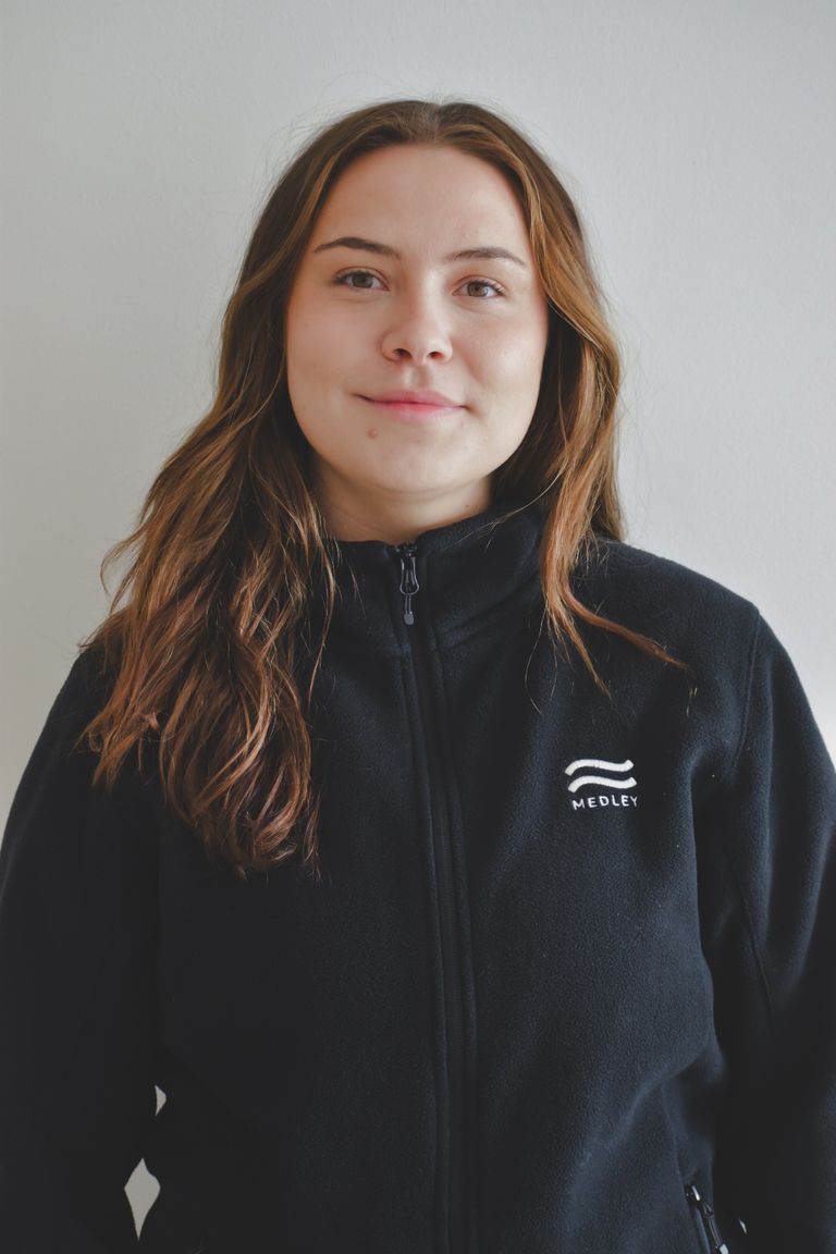 En ung kvinna med långt brunt hår, klädd i en svart jacka med dragkedja och MEDLEY-logotyp, står mot en vit bakgrund och ler mjukt mot kameran.