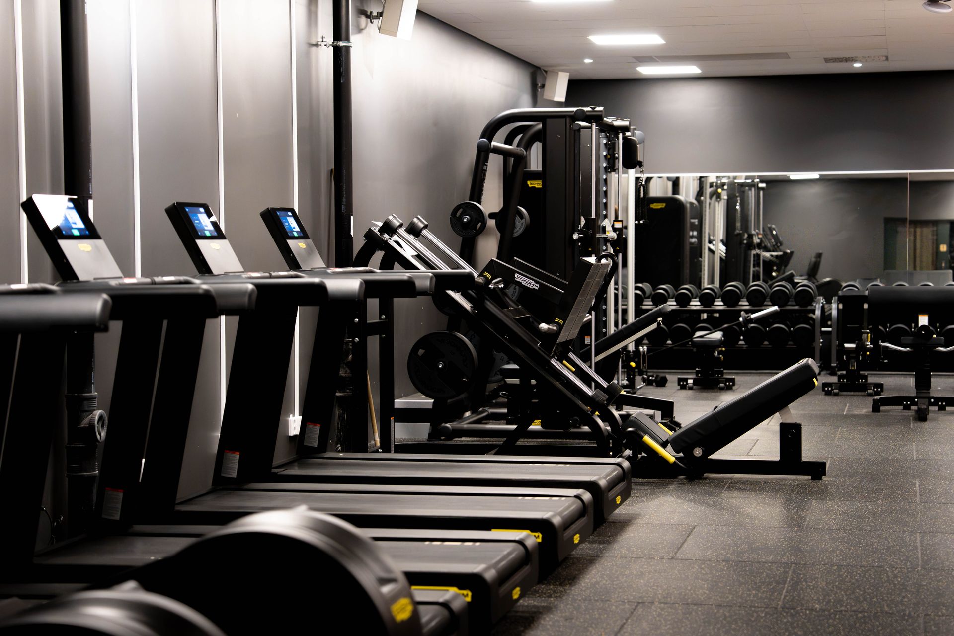 Interiörbild från ett modernt gym med en rad svarta löpband i förgrunden till vänster. I bakgrunden syns olika styrketräningsmaskiner, ett viktrack och en stor väggspegel som speglar utrustningen. Golvet har en mörk gummibeläggning.