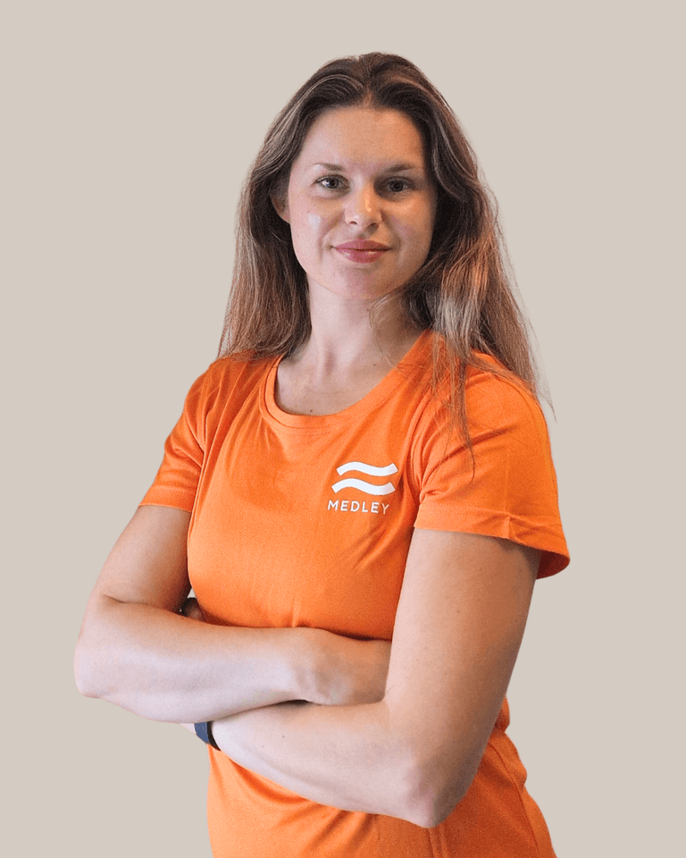 En kvinna med långt hår som bär en orange t-shirt med Medley-logotypen och står med armarna i kors.