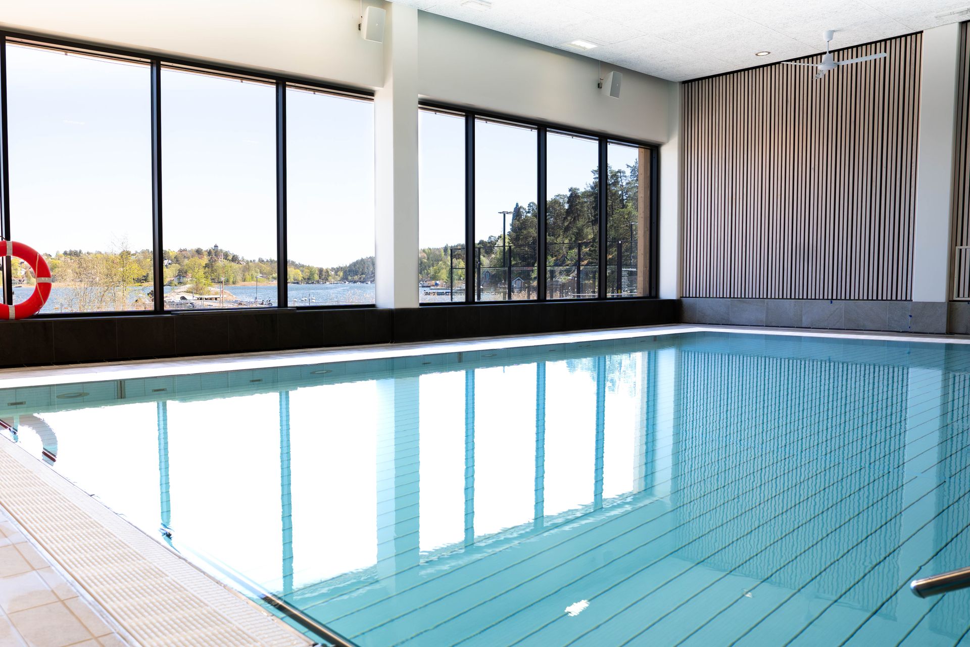 En inomhuspool med klart blått vatten, stora fönster från golv till tak och utsikt över en sjö och träd utanför. Solljuset strömmar in och speglas i vattnet. En röd livboj hänger på väggen bredvid fönstret.