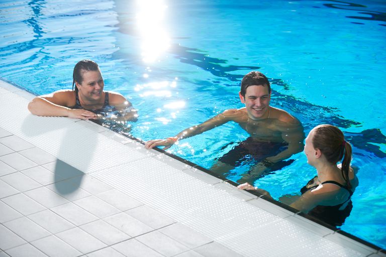 Tre personer i simkläder sitter i vattnet vid kanten på en simbassäng och samtalar glatt med varandra. Vattnet är ljusblått och på ytan skiner solen.