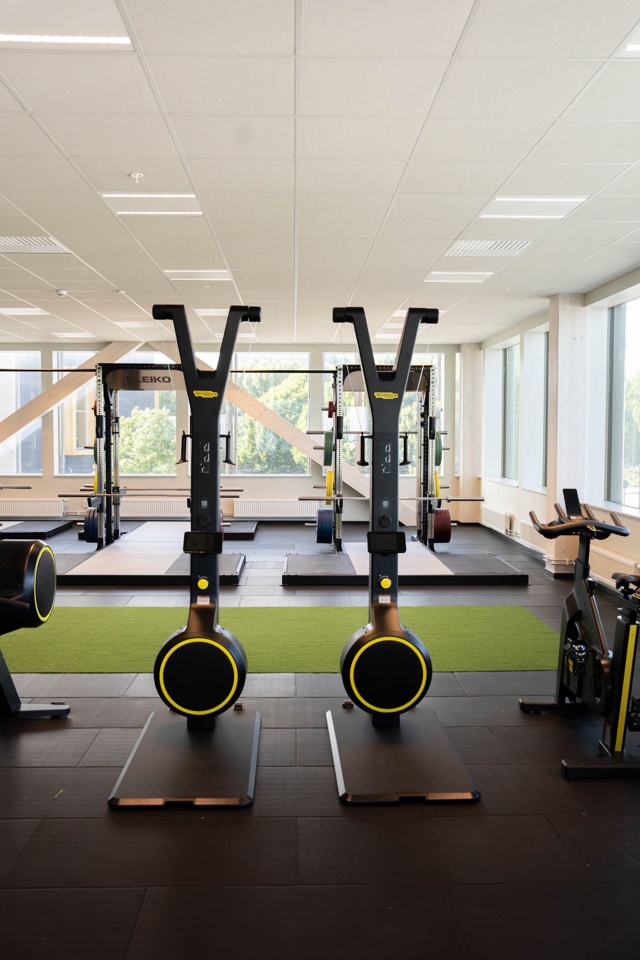 Ett ljust och modernt gym med stora fönster, där två skidergometer står i mitten, tyngdlyftningsplattformar längst bak och en motionscykel till höger.