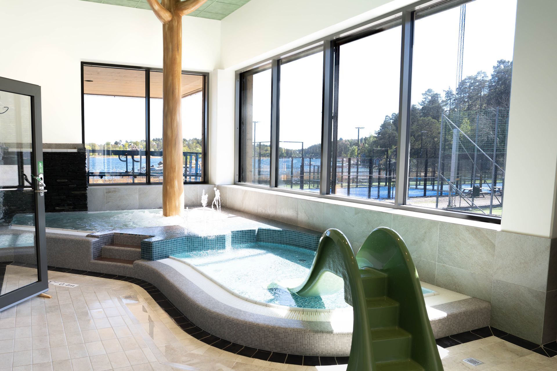 Inomhuspoolområde med en liten spapool, bubblande vatten, en grön rutschkana, stora fönster som släpper in naturligt ljus och utsikt över tennisbanor och träd utanför. Inredningen präglas av en guldfärgad trädkonstruktion i imiterat material.