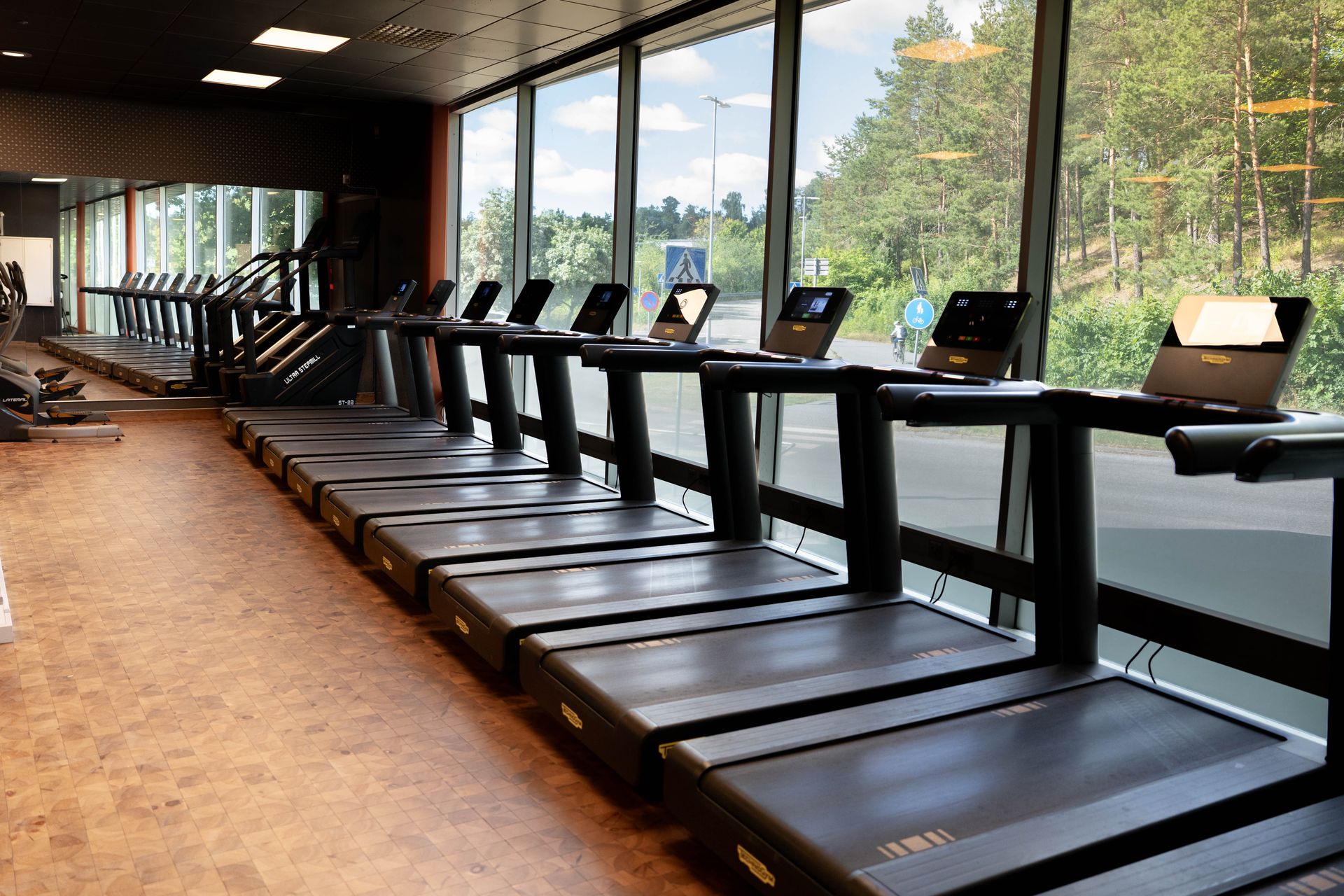 En rad löpband står uppställda i ett gym med stora fönster som vetter mot en grön, trädbevuxen utomhusmiljö. Gymmet har brunt golv och modern utrustning.